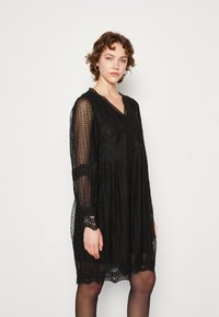 Schwarzes Kleid aus strukturierter Spitze mit langen Ärmeln, einem V-Ausschnitt und filigraner Stickerei entlang des Oberteils und des Saums. Lockerer Schnitt.