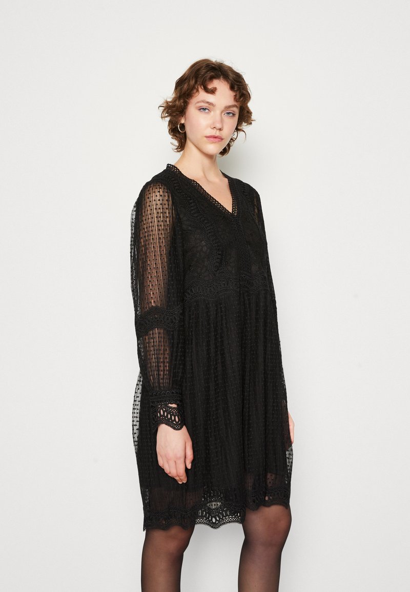Schwarzes Kleid aus strukturierter Spitze mit langen Ärmeln, einem V-Ausschnitt und filigraner Stickerei entlang des Oberteils und des Saums. Lockerer Schnitt.