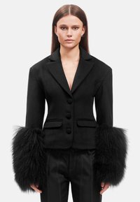 Blazer nero su misura con una silhouette aderente, caratterizzato da polsini in pelliccia testurizzata e bottoni frontali. Tasche su entrambi i lati.