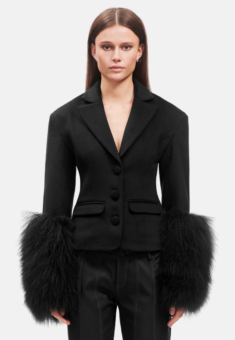 Blazer nero su misura con una silhouette aderente, caratterizzato da polsini in pelliccia testurizzata e bottoni frontali. Tasche su entrambi i lati.