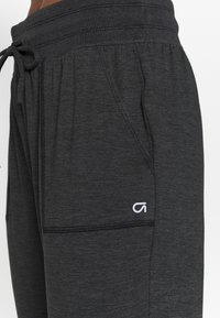 Tummanharmaat urheiluhousut, joissa on nauhavyötärö, sivutasku ja pieni valkoinen logo lähellä taskun saumaa.