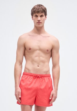 Coral swim trunks with an elastic waistband and drawstring. Smooth fabric, knee-length design, featuring a simple, solid color.