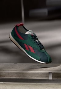 Green and navy Reebok sneaker with red stripes and black laces floating above a concrete surface in a dimly lit setting.