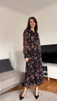 Hosszú fekete ruha színes virágmintával, áttetsző anyagból, gombos gallérral, hosszú ujjakkal és derékpánttal. Fekete magassarkúval viselve.