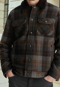 Uomo che indossa un giacca di lana a quadri marrone e grigia con collarino scuro e chiusura frontale con bottoni, con le mani nelle tasche dei pantaloni neri.