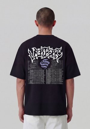 Man draagt een zwart T-shirt met witte grafische tekst en een paars slangontwerp op de achterkant, staand tegen een effen grijze achtergrond.