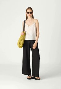 Top leveiro branco com detalhes bordados, calças largas pretas e uma mala texturizada verde oliva. A modelo usa óculos de sol pretos e sandálias.