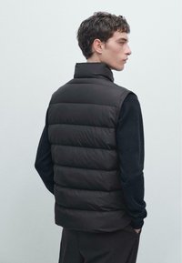 Gilet imbottito nero con cuciture orizzontali, colletto alto e design senza maniche, indossato sopra una camicia a maniche lunghe scura. Texture liscia.