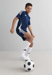 Zils sporta krekls ar baltajiem akcentiem un trim svītrām, apvienots ar atbilstošām šortiem, baltām zeķēm un baltām futbola kurpēm, stāv uz melnbalta futbola bumbas.