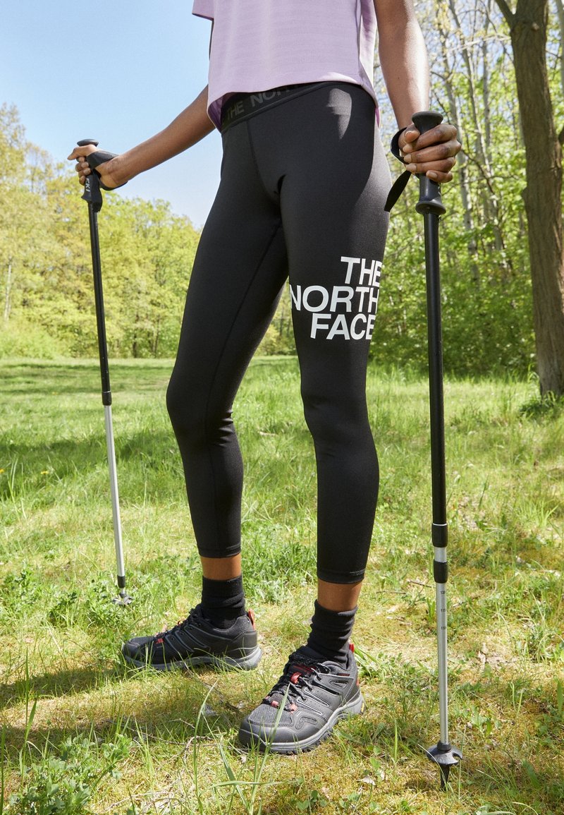 Černé legíny s logem "THE NORTH FACE" a vysokým pasem, v kombinaci s černými turistickými botami, držící trekingové hole v travnaté oblasti.