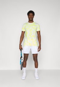 Lys gul og hvit mønster t-skjorte med korte ermer, kombinert med hvite shorts. Modellen holder en tennisracket og står mot en nøytral bakgrunn.