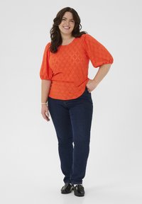 Ljusorange blus med strukturerat diamantmönster och puffiga ärmar, kombinerad med mörkblå skinny jeans och svarta skor.