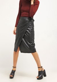 Svart fusk-läder omslagskjol med hög midja, diagonala framtill pleats och detalj med spänne. Matchas med kraftiga svarta klacksandaler.