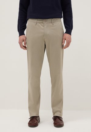 Uomo con pantaloni beige a taglio dritto, maglione blu scuro e scarpe stringate in pelle marrone, in piedi su uno sfondo chiaro e uniforme.