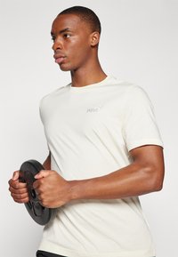 Tricou scurt cu mânecă, de culoare bej deschis, având un mic logo alb Puma. Persoana ține în mână o disc de greutate neagră, evidențiind mușchii puternici ai brațelor.
