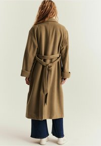 Olivgrüner Wollmischmantel mit tailliertem Gürtel, weiten Ärmeln und Reverskragen. Kombiniert mit dunkler Jeans und weißen Sneakers.