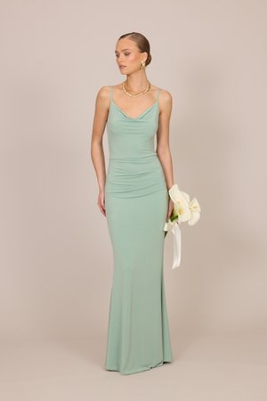 Γυναίκα με ανοιχτό πράσινο εφαρμοστό βραδινό φόρεμα κρατάει λευκές κάλλες με λευκή κορδέλα, φοράει χρυσά σκουλαρίκια και κολιέ, στέκεται σε μπεζ φόντο.