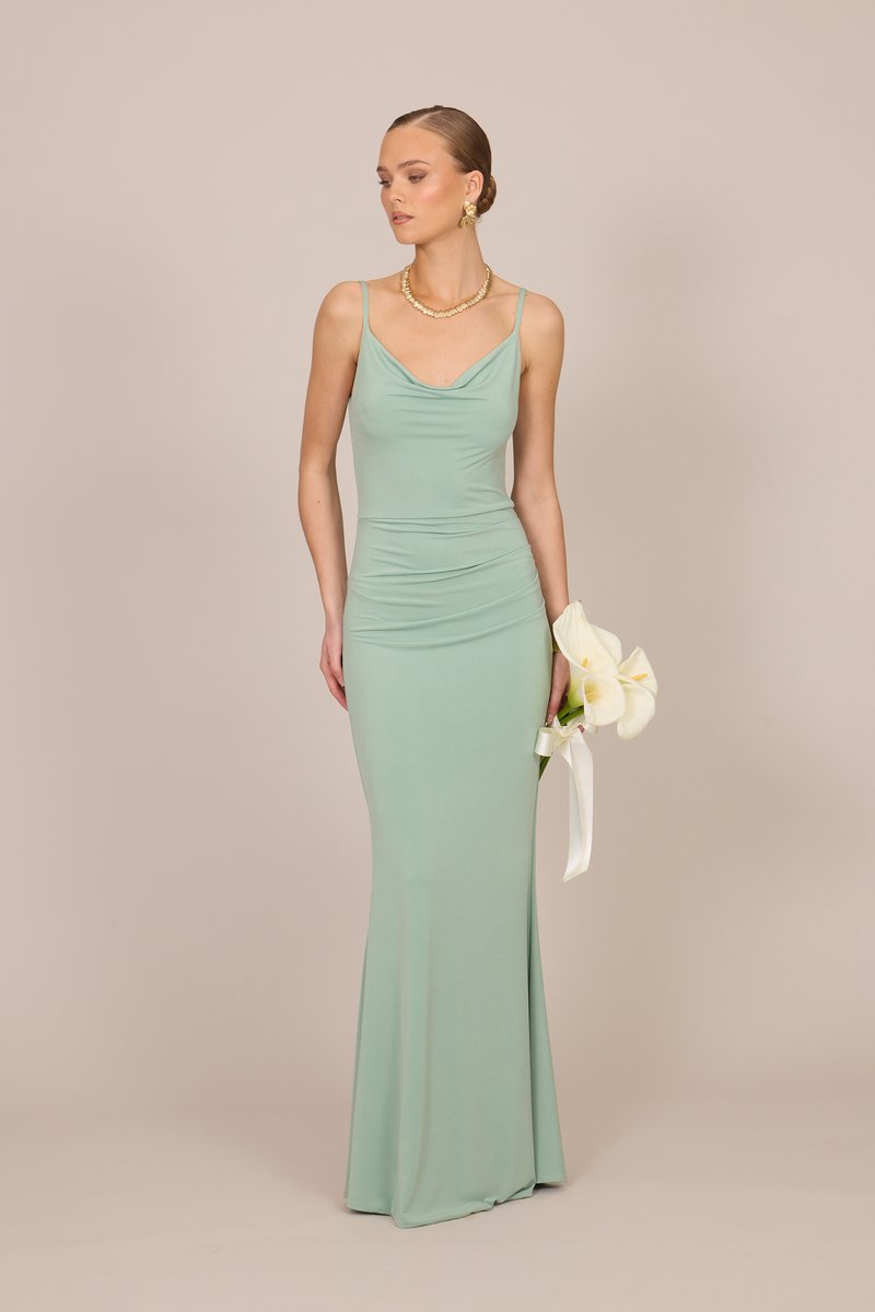 Γυναίκα με ανοιχτό πράσινο εφαρμοστό βραδινό φόρεμα κρατάει λευκές κάλλες με λευκή κορδέλα, φοράει χρυσά σκουλαρίκια και κολιέ, στέκεται σε μπεζ φόντο.