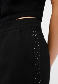 Pantalons ajustés noirs avec une texture lisse, présentant une ligne de petites studs scintillants sur le côté pour un détail supplémentaire.