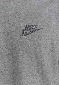 Šedá mikina vyrobená z texturované tkaniny, s pestrým třpytivým logem Nike na hrudi. V materiálu jsou jemné třpytky.