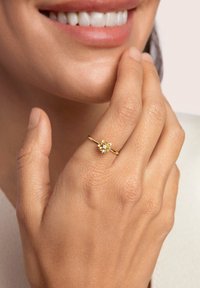 Anello d'oro con un design floreale dotato di una pietra centrale, montato su una sottile fascia. La mano mostra una pelle liscia e le unghie sono non lucidate.