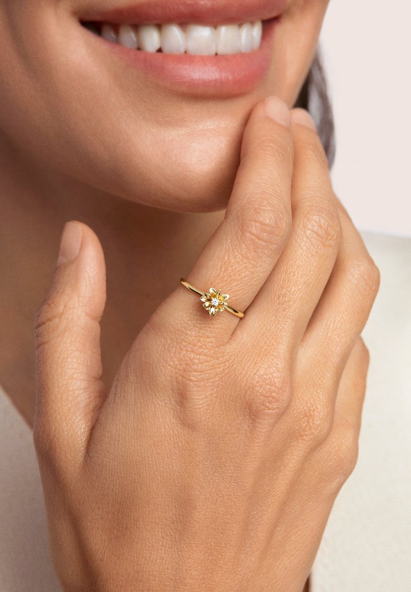 Anello d'oro con un design floreale dotato di una pietra centrale, montato su una sottile fascia. La mano mostra una pelle liscia e le unghie sono non lucidate.
