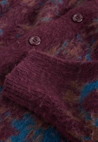 Détail d'un pull en tricot texturé bordeaux avec des accents bleus et marron, doté d'un poignet côtelé et d'une fermeture à bouton rond marron.