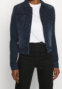 Džínová bunda tmavě modré barvy s přiléhavým střihem, zapínání na knoflíky, dvě náprsní kapsy a kontrastní šití. Nositá přes bílou košili.