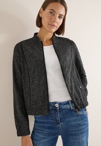 Svart och grå strukturerad jacka med en kortad silhuett, dragkedja framtill och subtila sömnadsdetaljer, kombinerad med en vit skjorta och blå jeans.