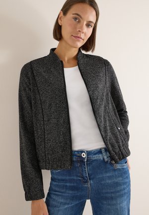 Schwarze und graue strukturierte Jacke mit einer gekürzten Silhouette, Reißverschluss vorne und dezenten Nähdetails, kombiniert mit einem weißen Shirt und blauen Jeans.