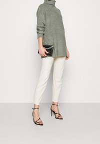 Jersey de cuello alto verde oversize, pantalones crema ajustados, tacones negros con tiras y un bolso negro de mano con un acento de cadena.