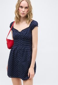 Νεαρή γυναίκα με ξανθά μαλλιά μέχρι τους ώμους, ντυμένη με μπλε σκούρο φόρεμα με πουά και κρατώντας μικρή κόκκινη τσάντα ώμου.