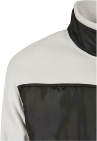Veste en polaire blanche avec un col noir haut et un panneau noir horizontal au niveau de la poitrine et des épaules.