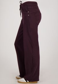 Zijaanzicht van een persoon in donkerpaarse wijde broek met een trekkoord in de taille en witte sneakers met zwarte strepen.