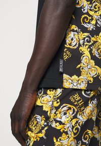 Dunkelhäutiger Arm neben schwarzer Kleidung mit aufwendigen gelben und weißen barocken Mustern und Text-Tags "Versace Jeans Couture".