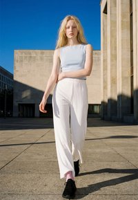 Jeune femme aux longs cheveux blonds portant un haut bleu clair sans manches, un pantalon blanc et des baskets noires, marchant en extérieur près de bâtiments en béton.