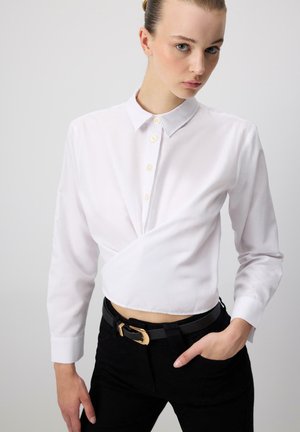 Młoda osoba ubrana w białą, długorękawową, krótką koszulę na guziki oraz czarne spodnie z czarnym paskiem z złotą klamrą.