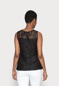 Donna con capelli corti e ricci, che indossa un top in pizzo nero senza maniche e pantaloni bianchi, vista di spalle su uno sfondo neutro.