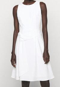 Bílá bezrámová šaty s těsným korzetem a splývavou sukní, s nařaseným detailem v pase. Hladká látka s jemným leskem.