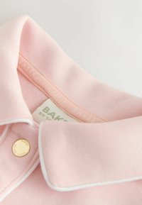 Weiches, rosa Kleidungsstück mit Kragen, weißer Paspelierung und einem goldenen Knopf mit der Aufschrift "Ted Baker" am Kragen.