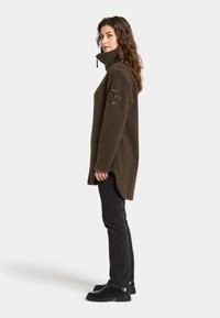 Übergroßer Fleece-Pullover in dunkelolivgrün mit hohem Kragen, strukturiertem Stoff und Lederpatch-Detail am Ärmel. Schwarze Hose und Schuhe.