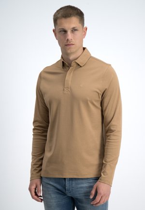 Jonge man die een beige polo met lange mouwen en blauwe spijkerbroek draagt, staat voor een effen witte achtergrond en kijkt naar links.