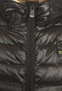 Sort puffjakke med glat finish, der har en frontlynlås, hætte og en lomme med en gul logoaccent. Taktile quiltede mønster.