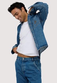 Jeansjacka i medelblått med knäppning, två framfickor och avslappnad passform, bärs över en vit t-shirt och matchande jeansbyxor.