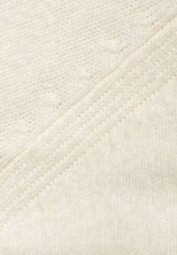 Strickstoff in Cremefarbe mit einer weichen Textur. Verfügt über diagonale Rippen und ein Muster aus kleinen erhabenen Formen, die sich vor einem glatten Hintergrund abheben.