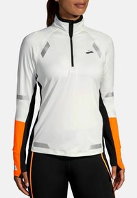 Maglietta atletica a maniche lunghe da donna, bianca con dettagli neri e arancioni, design a zip quarti, dettagli riflettenti, materiale sintetico elasticizzato.