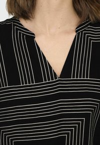 Top nero con scollo a V decorato da un motivo geometrico di linee bianche, che creano rettangoli e strisce verticali; il tessuto appare liscio con una leggera texture.