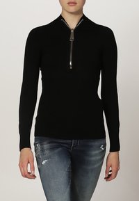 Czarny sweter o prążkowanej strukturze z wysokim kołnierzem i srebrnym zapięciem na zamek, zestawiony z podartymi niebieskimi dżinsami.