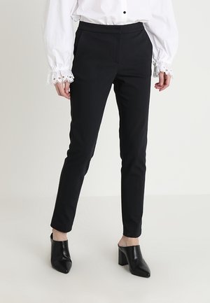 Pantalones negros ajustados con una textura suave, que cuentan con una cintura a media altura y un detalle frontal mínimo, combinados con tacones altos negros de punta abierta.