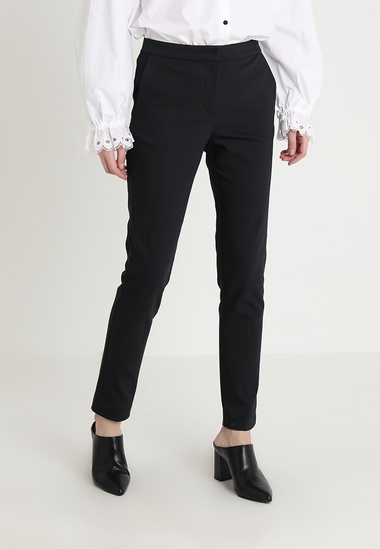 Czarne dopasowane spodnie o gładkiej teksturze, z talią o średnim wzroście i minimalnymi detalami z przodu, w połączeniu z czarnymi, odkrytymi szpilkami.
